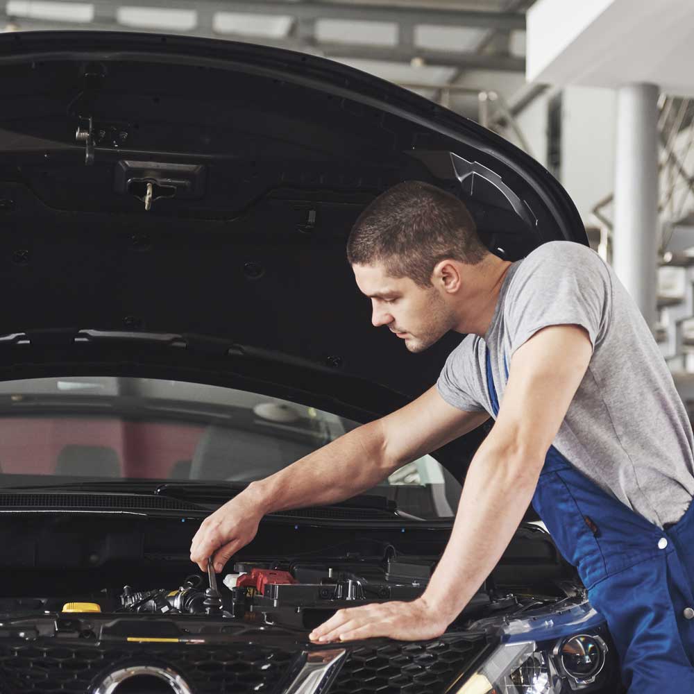A mechanic in a grey shirt and blue overalls leaning over a car's open hood.